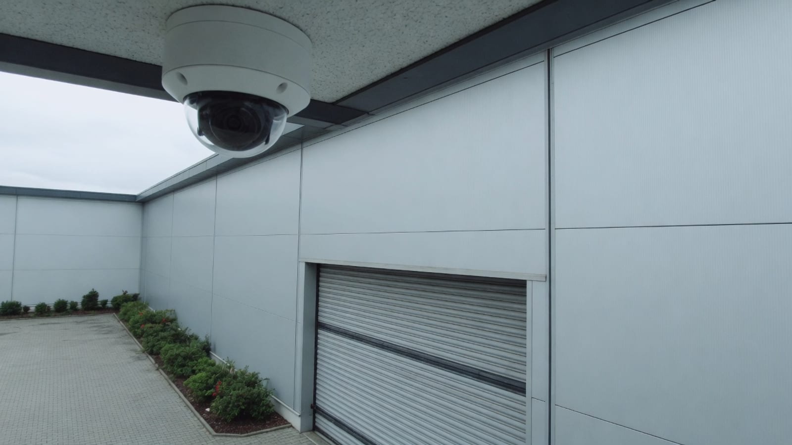 Kamera kopułkowa CCTV zamontowana pod okapem hali magazynowej, obok zamkniętej bramy rolowanej.